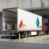 A large commercial truck parked at a loading dock, displaying diamond-shaped hazmat placards that indicate the transport of hazardous materials.